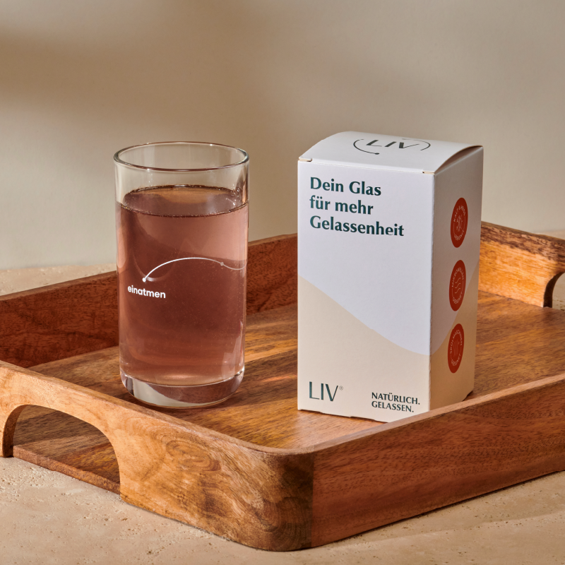 Ein Glas mit rosa Flüssigkeit und seine Umverpackung stehen auf einem Tablet aus Holz.