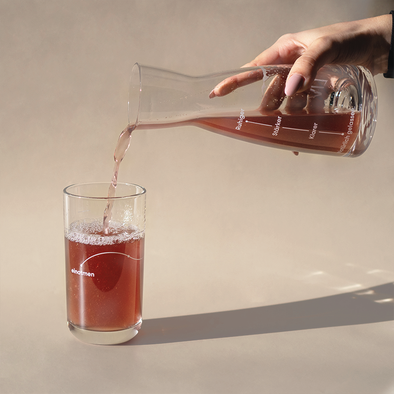 and gießt mit einer Kristallglas-Karaffe rosa Wasser in ein Glas. Das Wasser schäumt leicht auf, während der Hintergrund vom Bild beige ist.
